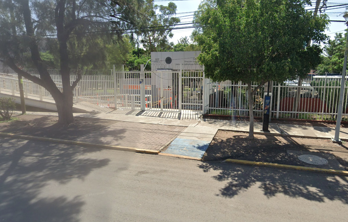 Fotografía en la que se muestra la entrada del plantel, conformada por una reja de color blanco que rodea las instalaciones. En medio se encuentra el acceso y frente a ella una rampa.